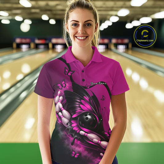 Female model at a bowling lane wearing a pink and black bowling polo shirt with butterfly artwork embracing a bowling ball and pins.