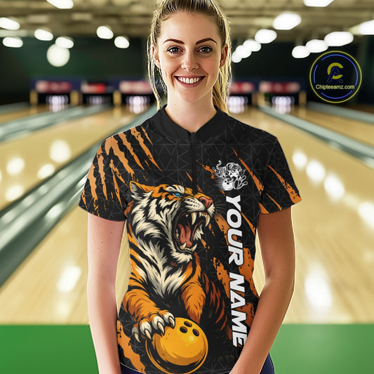 Front view of a female model at a bowling lane wearing a black and yellow bowling quarter-zip shirt featuring a roaring tiger holding a yellow bowling ball with orange claw-stripe graphics and a geometric background pattern.