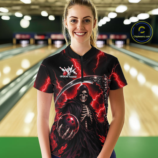 A woman standing in a bowling alley wearing a black short-sleeve bowling shirt with a red and black grim reaper graphic, featuring a bowling ball and skeleton design.