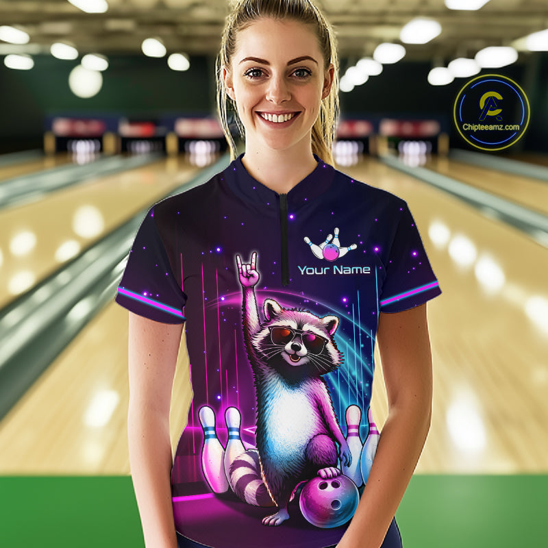 A woman stands in a bowling alley wearing a custom short-sleeve polo shirt with a colorful graphic of a raccoon holding a bowling ball and pins, with the text 'Your Name' and a bowling pin logo on the chest. 