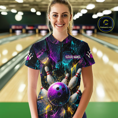 A woman standing in a bowling alley wearing a short-sleeve bowling-themed t-shirt with a colorful cosmic bowling pin and ball graphic. The shirt features the text 'YOUR NAME'