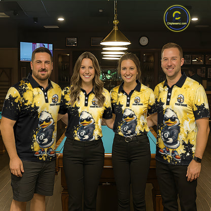 Four people standing together in a dimly lit indoor venue, wearing matching yellow and black polo shirts with a cartoon duck graphic and the text 'YOUR NAME'