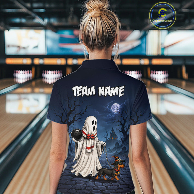 A woman viewed from behind wearing a navy blue bowling-themed polo shirt with a Halloween graphic of a ghost, a dog, and the text 'TEAM NAME'. The background shows a blurred bowling alley interior.