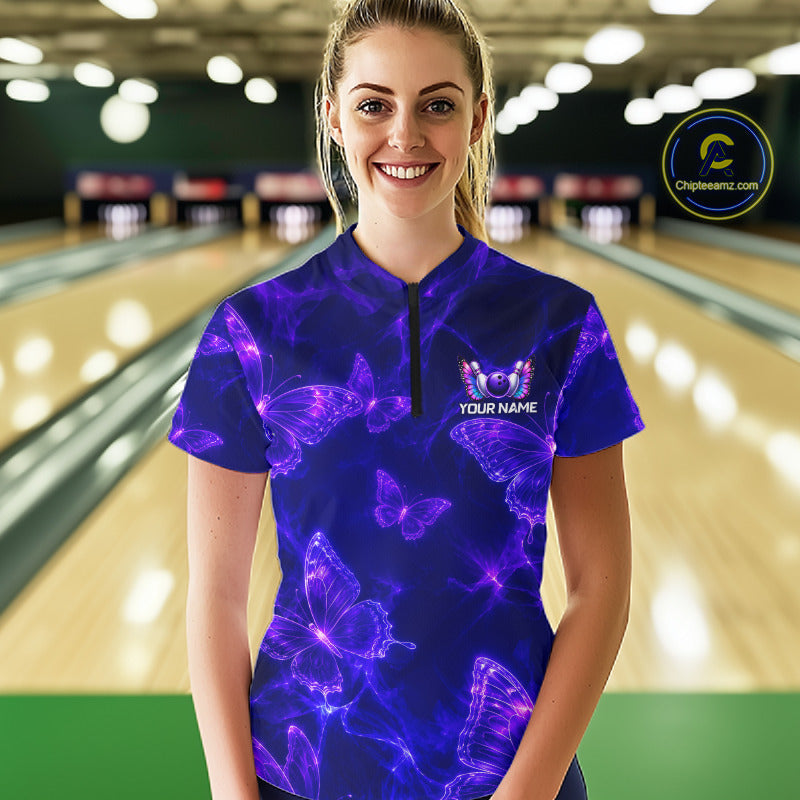 A woman wearing a purple and blue bowling-themed short-sleeve polo shirt with glowing butterfly graphics, standing in a bowling alley. The shirt features a black zipper at the collar and a customizable chest graphic that reads 'YOUR NAME'.