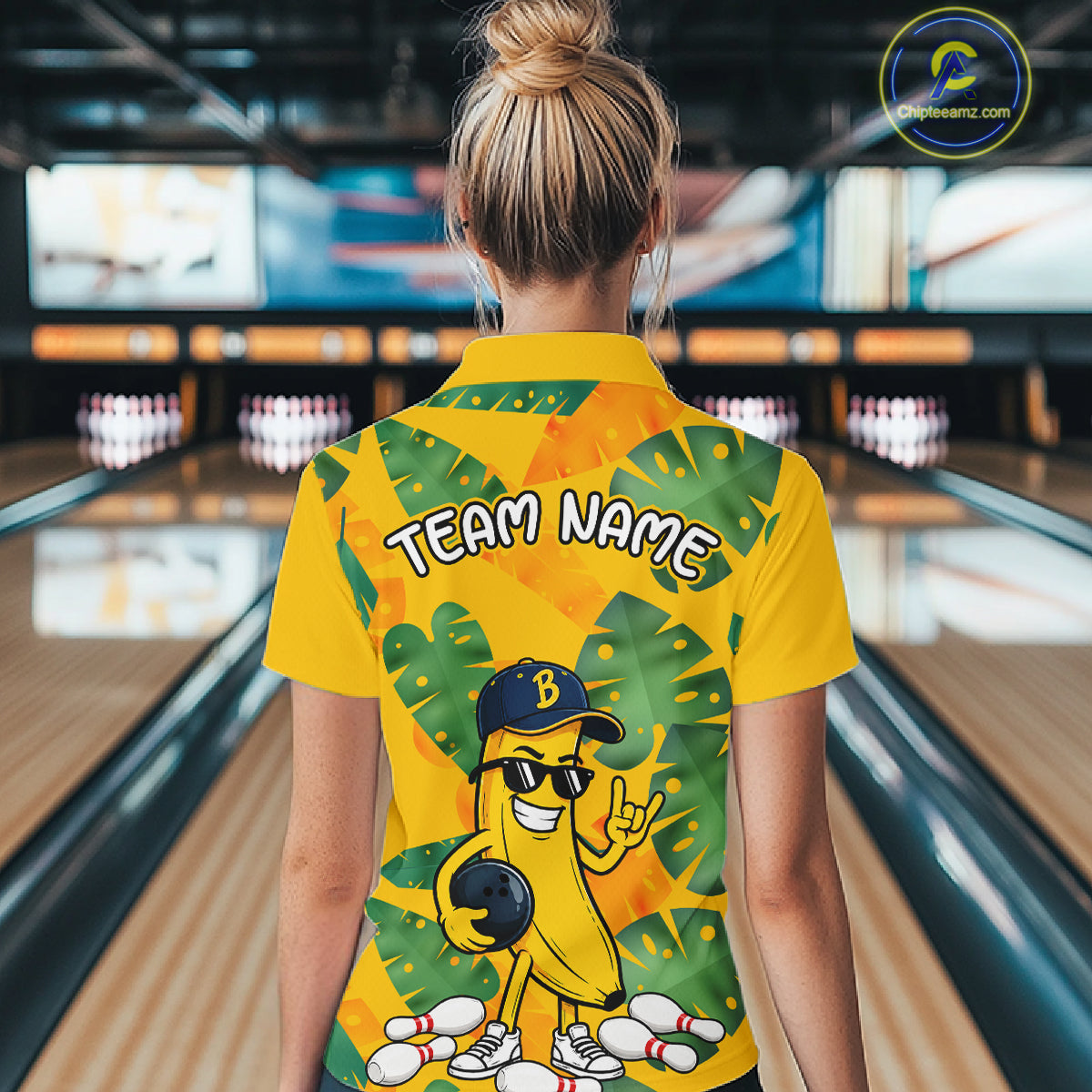 Woman in a bowling alley wearing a bright yellow tropical polo shirt with green leaf patterns, custom team name text, and a smiling banana mascot holding a bowling ball.