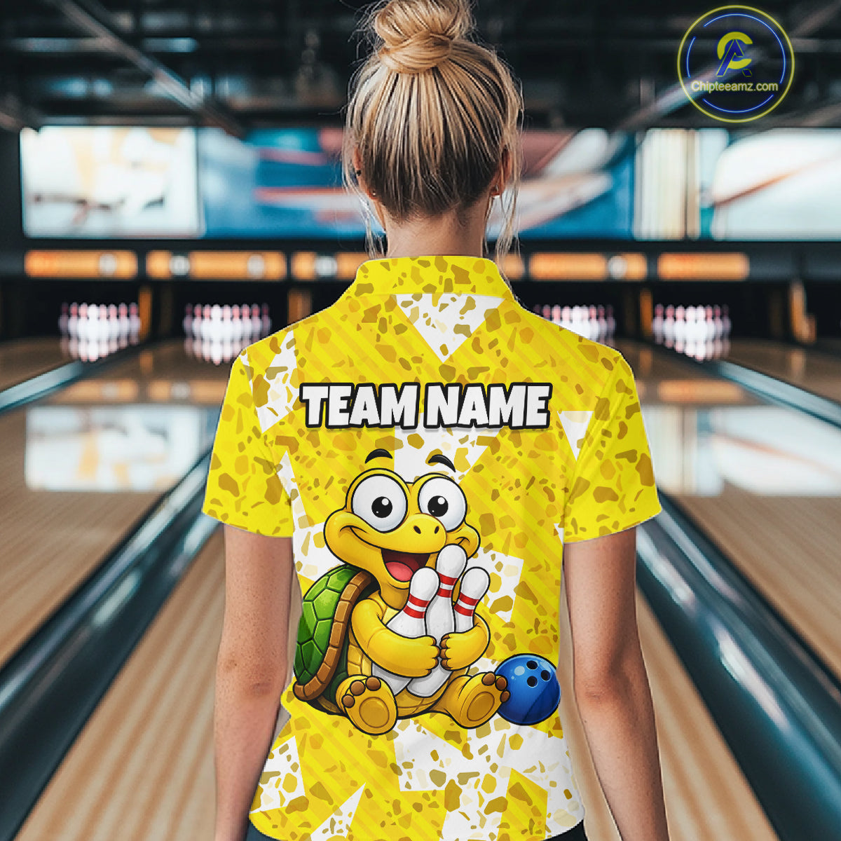 Woman wearing a yellow bowling-themed shirt with a cartoon turtle holding a bowling ball in a bowling alley.