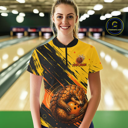 A woman stands in a bowling alley wearing a short-sleeve bowling shirt with a yellow and black paint-splatter design, a large pangolin graphic, and a bowling pin logo on the chest. 