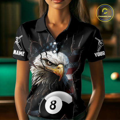 Woman wearing a black polo shirt with an eagle and pool ball design, holding a pool cue in a billiards setting.