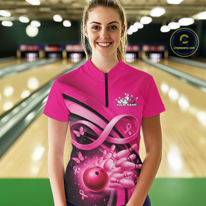 Female model wearing a pink bowling quarter zip with pink ribbon artwork, delicate butterflies, and bowling graphics at the bowling lane.