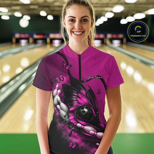 Female model wearing a pink and black bowling quarter-zip shirt at the bowling lane with butterfly artwork surrounding a bowling ball and pins.