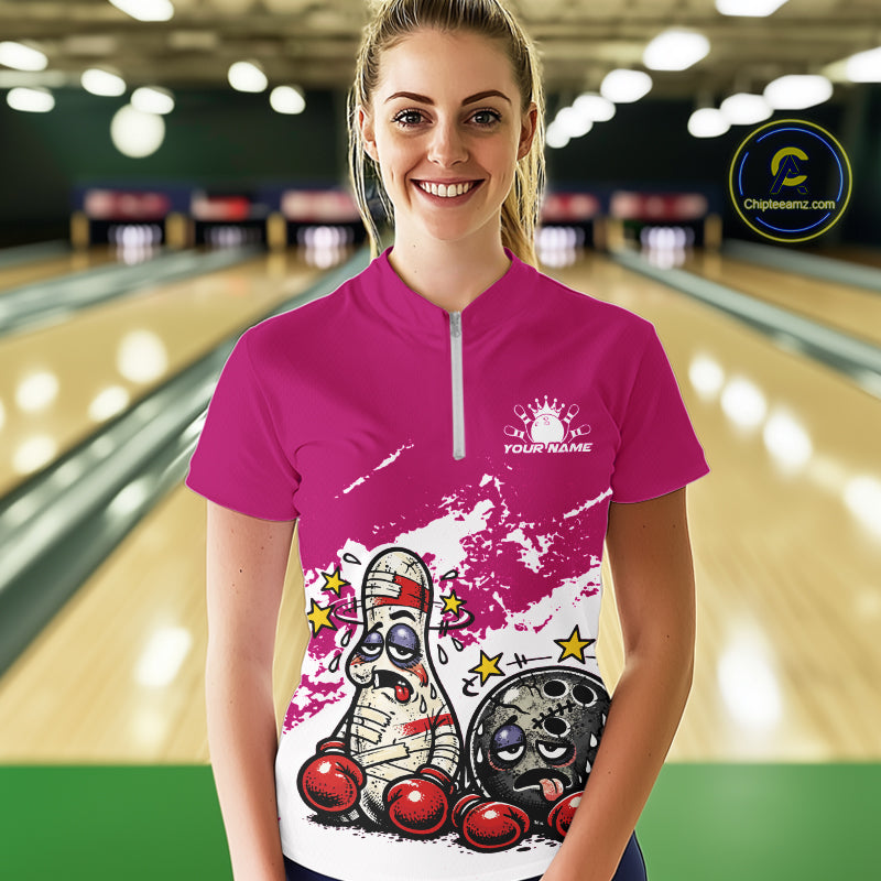 Female model at a bowling lane wearing a pink and white quarter-zip shirt, cartoon boxing bowling ball and pin artwork with fun sporty vibe.