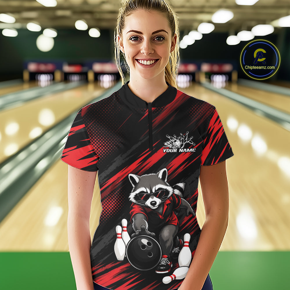 A woman stands in a bowling alley wearing a custom short-sleeve bowling quarter zip  shirt. The shirt features a black and red abstract background with a cartoon raccoon holding a bowling ball and pins, and a white text placeholder 'YOUR NAME'.