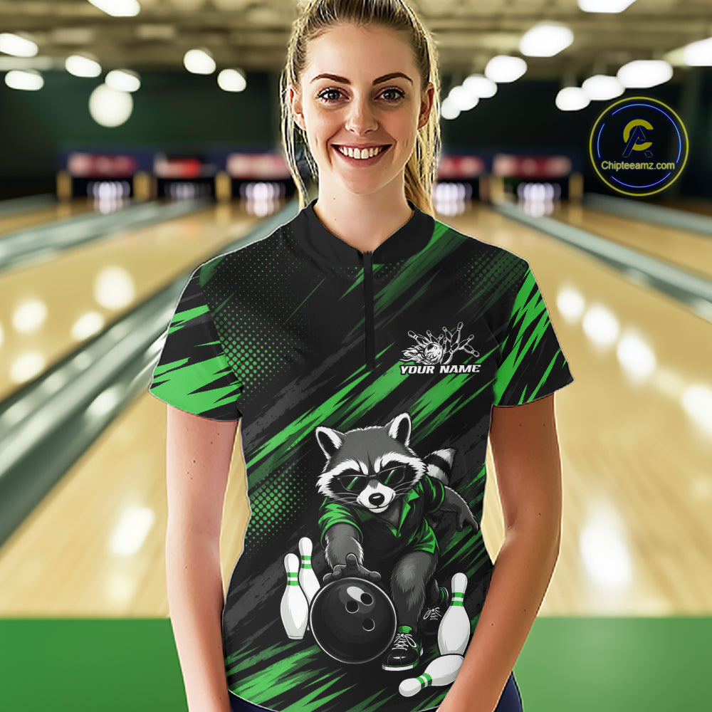 A woman stands in a bowling alley wearing a custom short-sleeve bowling quarter zip shirt. The shirt features a black and green abstract pattern with a cartoon raccoon holding a bowling ball and pins, and a mock logo that reads 'YOUR NAME'.