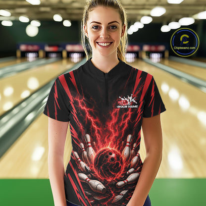 A woman wearing a black quarter zip  bowling jersey with a red and black lightning graphic and bowling pins holding a bowling ball, standing in a bowling alley.