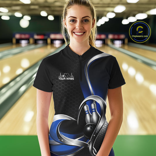 Female model in a bowling alley wearing a black and blue quarter zip shirt, showing a honeycomb pattern and heart motif surrounding a bowling ball and pins.