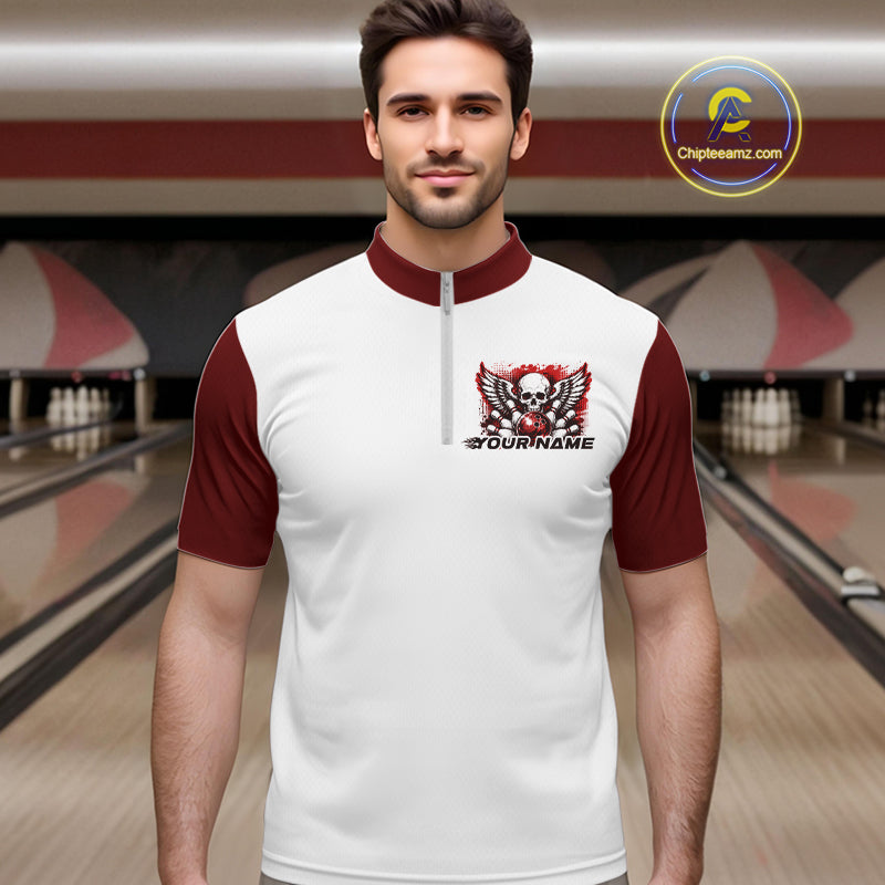 A man stands in a bowling alley wearing a bowling shirt with quarter zip collar, red skull-themed bowling graphic with black wings, pins, bowling ball