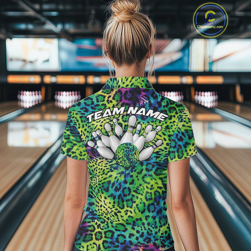 Back view of a woman standing at a bowling alley, wearing a green, blue, and black leopard-print bowling polo with “Team Name” and bowling pins on the back.