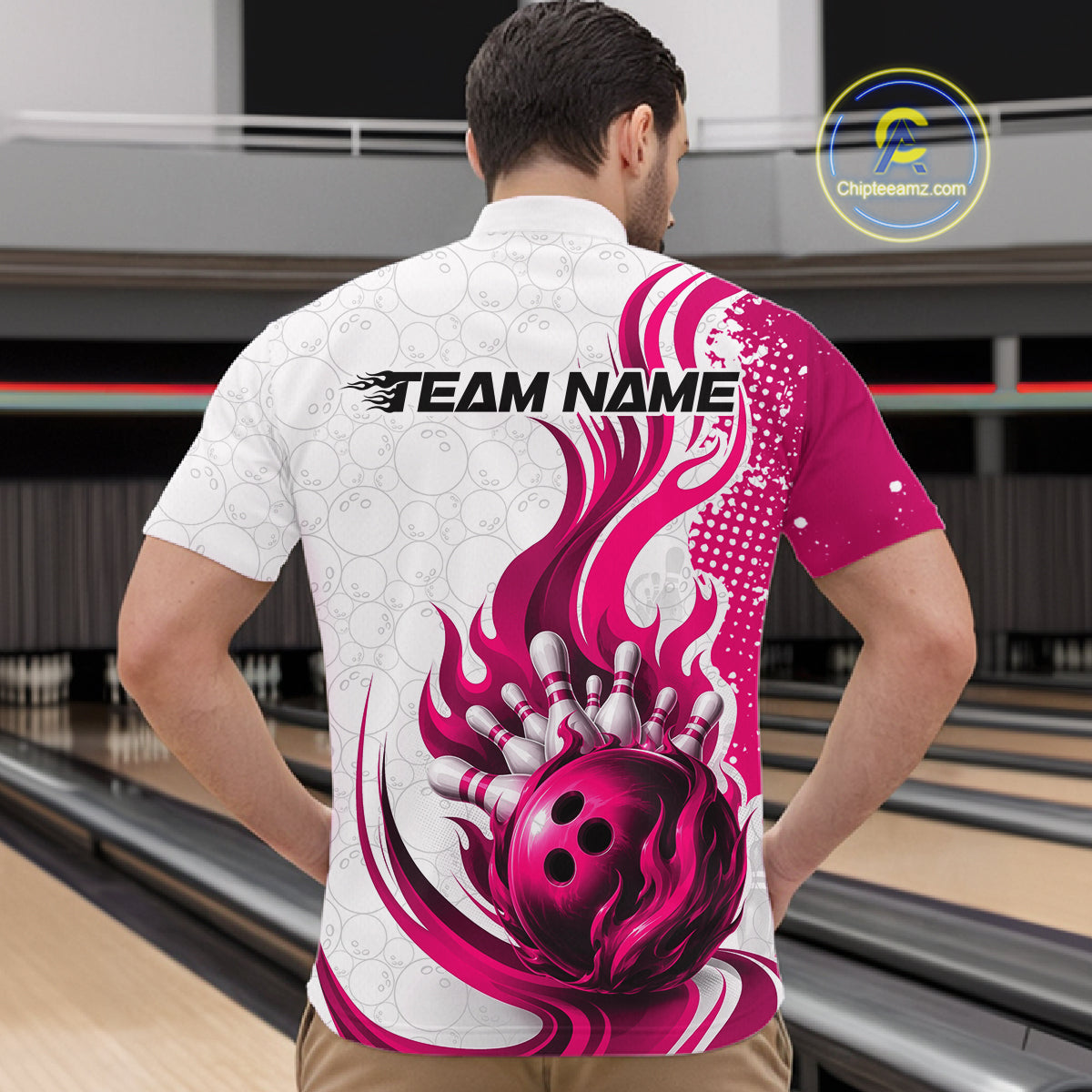 A man viewed from behind wearing a short-sleeve bowling-themed shirt with a white and magenta flame-and-ball graphic, standing in a bowling alley. The shirt has 'TEAM NAME' printed across the upper back