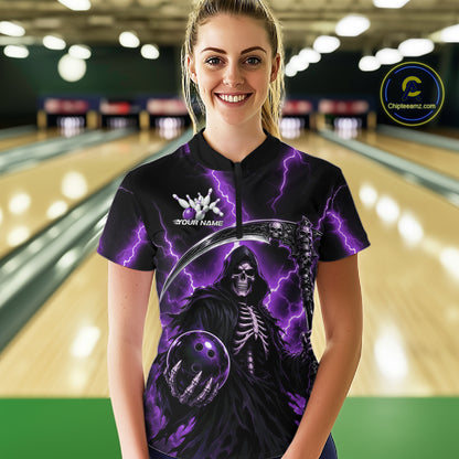 A woman stands in a bowling alley wearing a black short-sleeve polo shirt with a purple lightning and grim reaper graphic. 