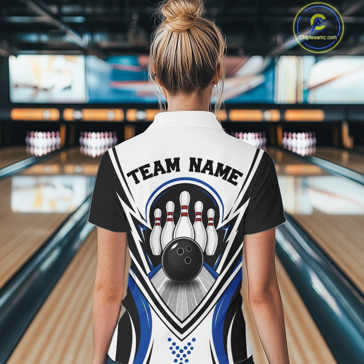 Back view of a female model inside a bowling center wearing a white, black, and blue bowling quarter-zip shirt with bowling-themed graphics of a bowling ball and pins.