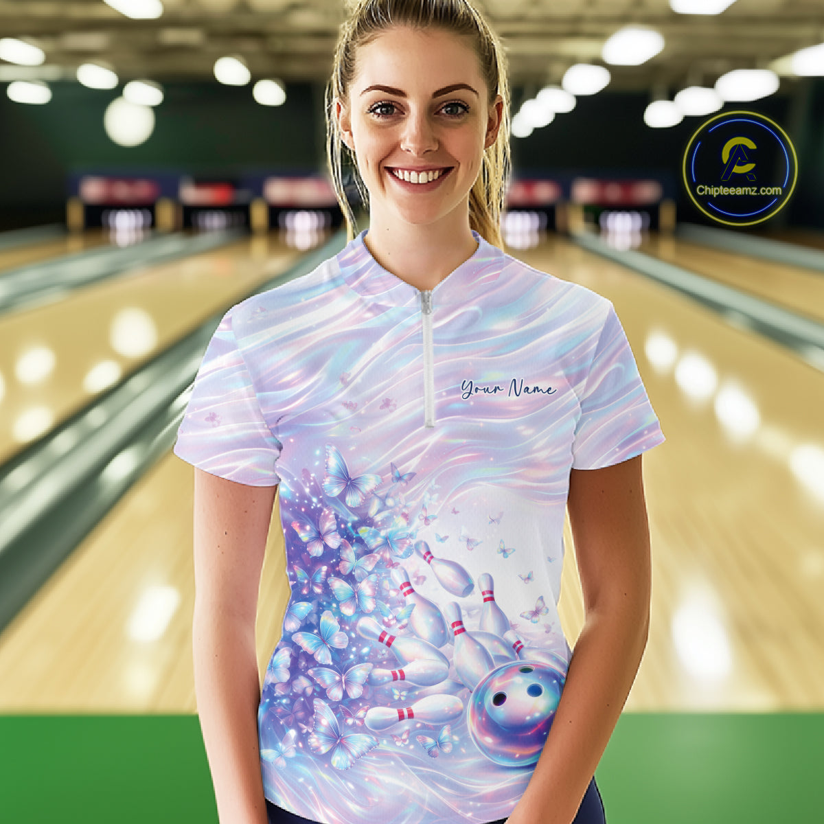Female model wearing a bowling quarter zip with pastel purple and pink holographic butterfly graphics and bowling accents, front view at the bowling lane