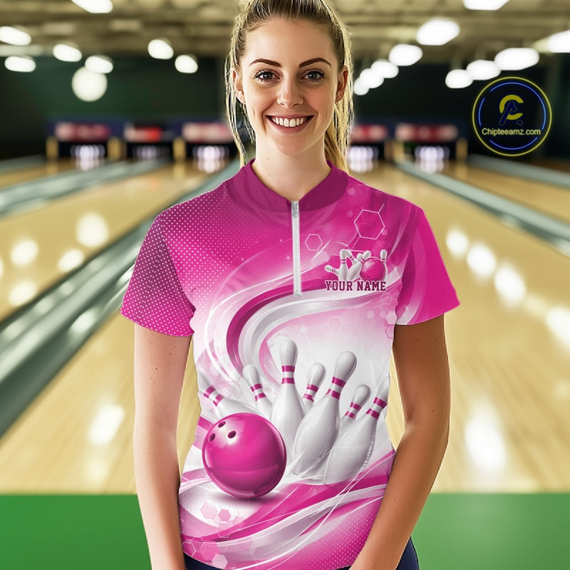 Female model standing in a bowling lane wearing a white and pink bowling quarter-zip shirt featuring abstract wave lines and a glossy bowling ball striking pins on the front.