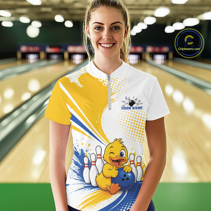 A woman stands in a bowling alley wearing a custom short-sleeve bowling quarter zip shirt. The shirt features a white upper chest and collar, a yellow and blue graphic splash, and a cartoon yellow duck holding a blue bowling ball next to white bowling pins.