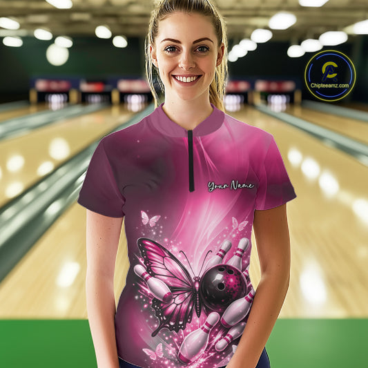 Female model shown from the front in a bowling alley wearing a pink quarter-zip shirt featuring butterfly artwork, a bowling ball, and pins.