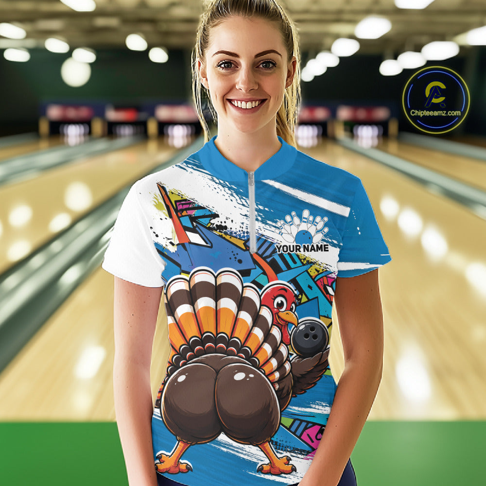 Front view of a happy female model in a bowling alley wearing a blue custom quarter zip bowling shirt with a funny graffiti turkey holding a bowling ball, paint splatter accents, and a personalized name.