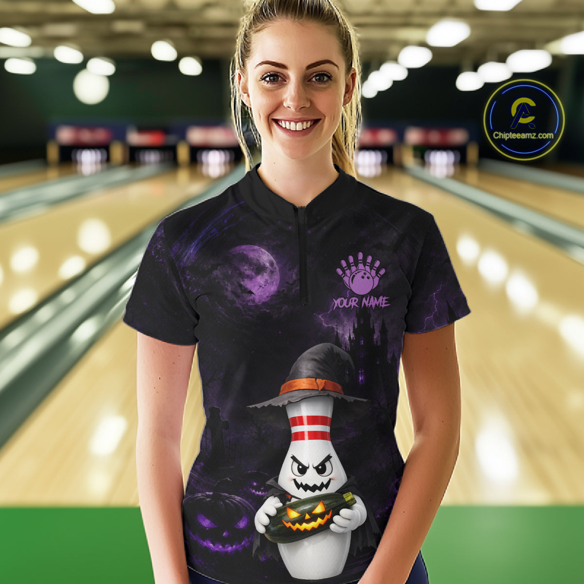 A woman stands in a bowling alley wearing a black short-sleeve polo shirt with a Halloween-themed graphic print. The shirt features a purple moon and stormy sky background, a cartoon ghost holding a jack-o'-lantern, and a purple placeholder text reading 'YOUR NAME'.