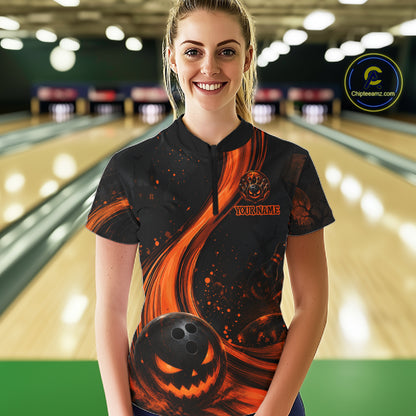 A woman stands in a bowling alley wearing a black short-sleeve bowling shirt with a Halloween-themed orange and black graphic print featuring a jack-o'-lantern and swirls. The background shows blurred bowling lanes