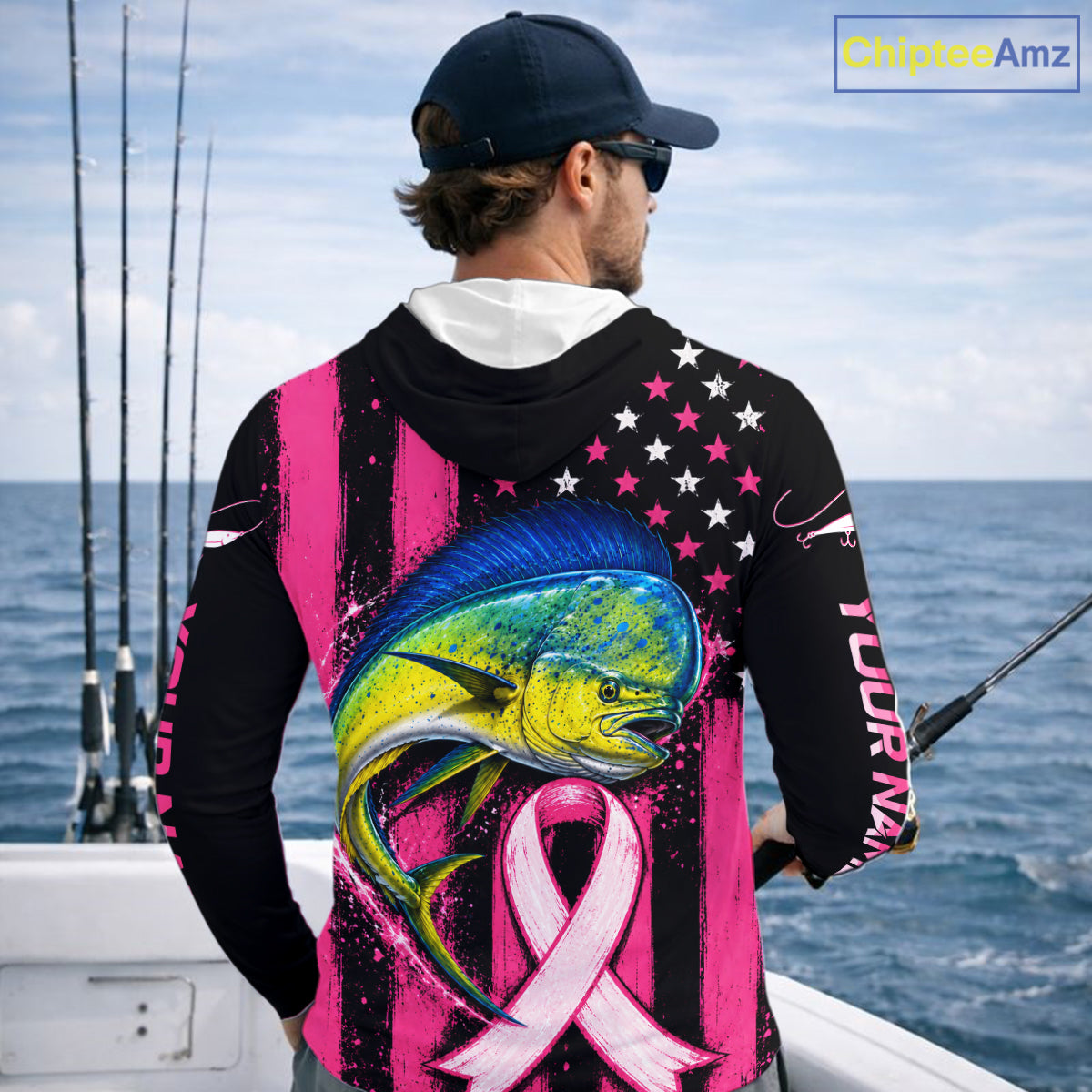 A man on a boat wearing a black and pink fishing hoodie with a large marlin graphic, pink awareness ribbon, and American flag pattern on the back. The hoodie has 'YOUR NAME' printed on the sleeve.