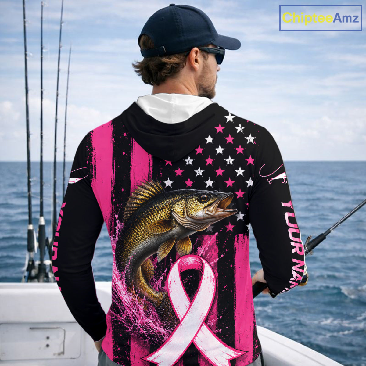 A man wearing a black and pink long-sleeve hoodie with a walleye fish graphic and pink ribbon design on the back, standing on a boat with fishing rods and the ocean in the background.