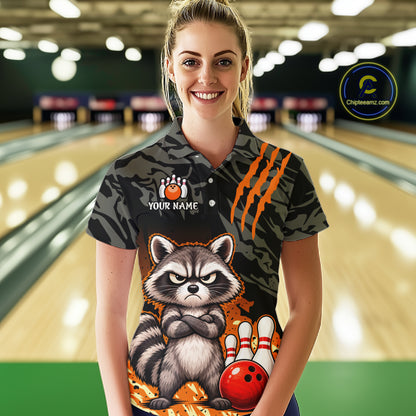 A woman stands in a bowling alley wearing a custom short-sleeve polo shirt with a raccoon graphic, orange claw marks, and bowling pins and balls. The shirt has a camouflage pattern on the sleeves and upper chest, and a colorful raccoon design on the lower front.