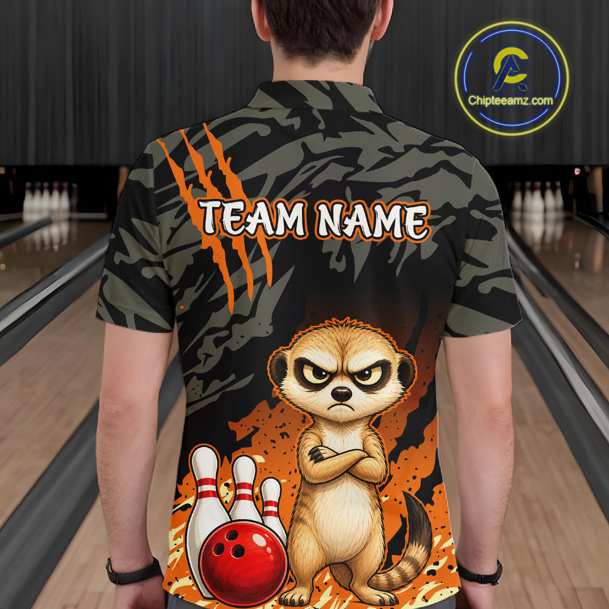 A person shown from behind wearing a short-sleeve bowling-themed shirt with a dark camouflage pattern, orange claw marks, the text 'TEAM NAME', and a cartoon meerkat standing in front of bowling pins and a red bowling ball. The background is a blurred bowling alley.