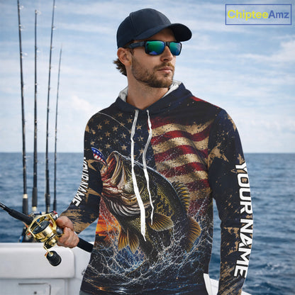 Man on a boat wearing a hooded fishing shirt with a fish design and American flag, holding a fishing rod.